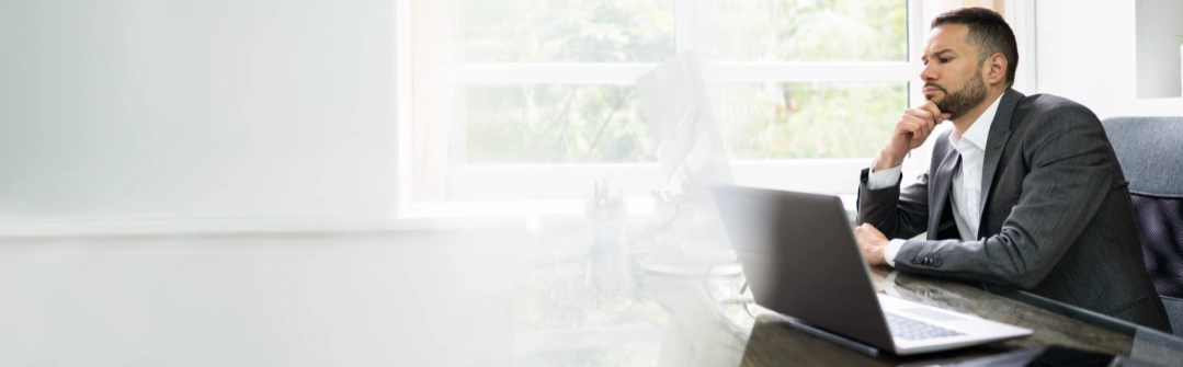 businessman at desk behind computer thinking
